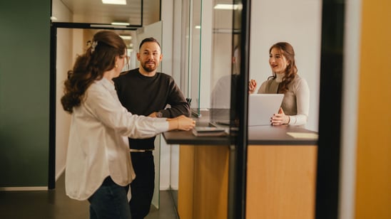 A man and a woman stand at the counter of a public authority. Behind the counter is a public authority employee, next to her a notebook on which she wants to show the two of them something in front of the counter.