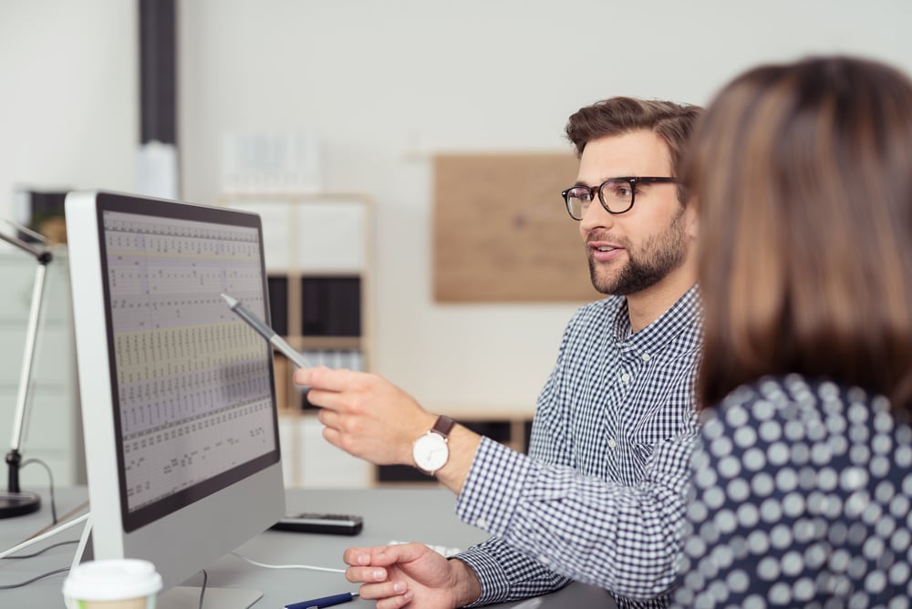 Proficient young male employee with eyeglasses and checkered shirt, explaining a business analysis displayed on the monitor of a desktop PC to his female colleague, in the interior of a modern office Proficient young male employee with eyeglasses and checkered shirt, explaining a business analysis displayed on the monitor of a desktop PC to his female colleague, in the interior of a modern office