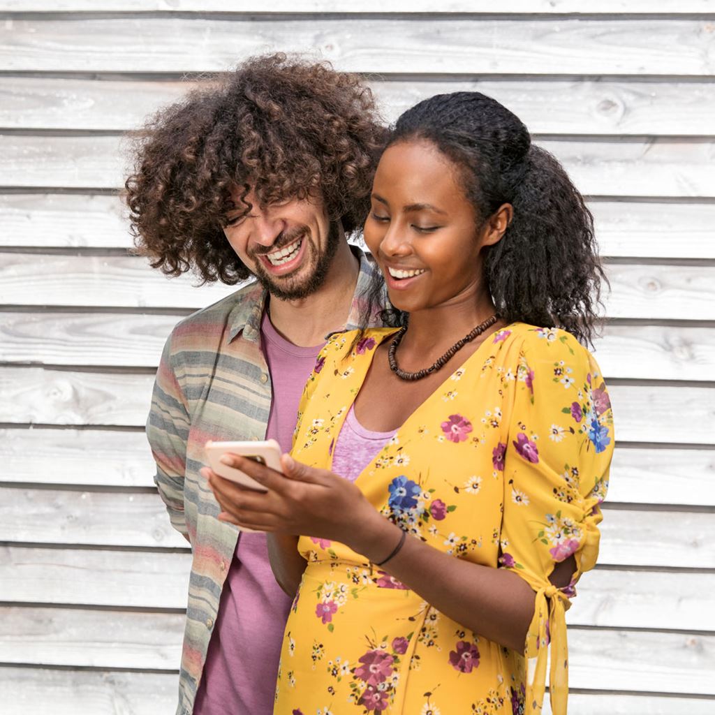 Due persone che ridono, una con un vestito giallo e l'altra con una maglietta a righe, guardano insieme uno smartphone: un simbolo dell'uso condiviso delle app per il voto elettronico.