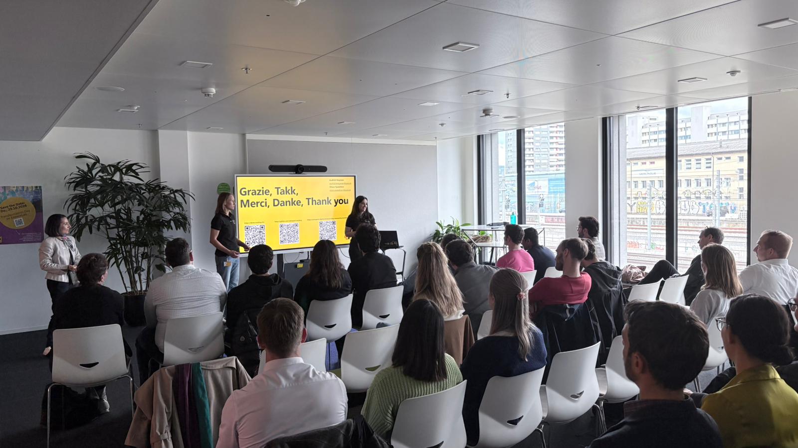 An audience seated in a room listens to a presentation; at the front, a group stands next to a screen showing a multilingual thank-you message and QR codes.