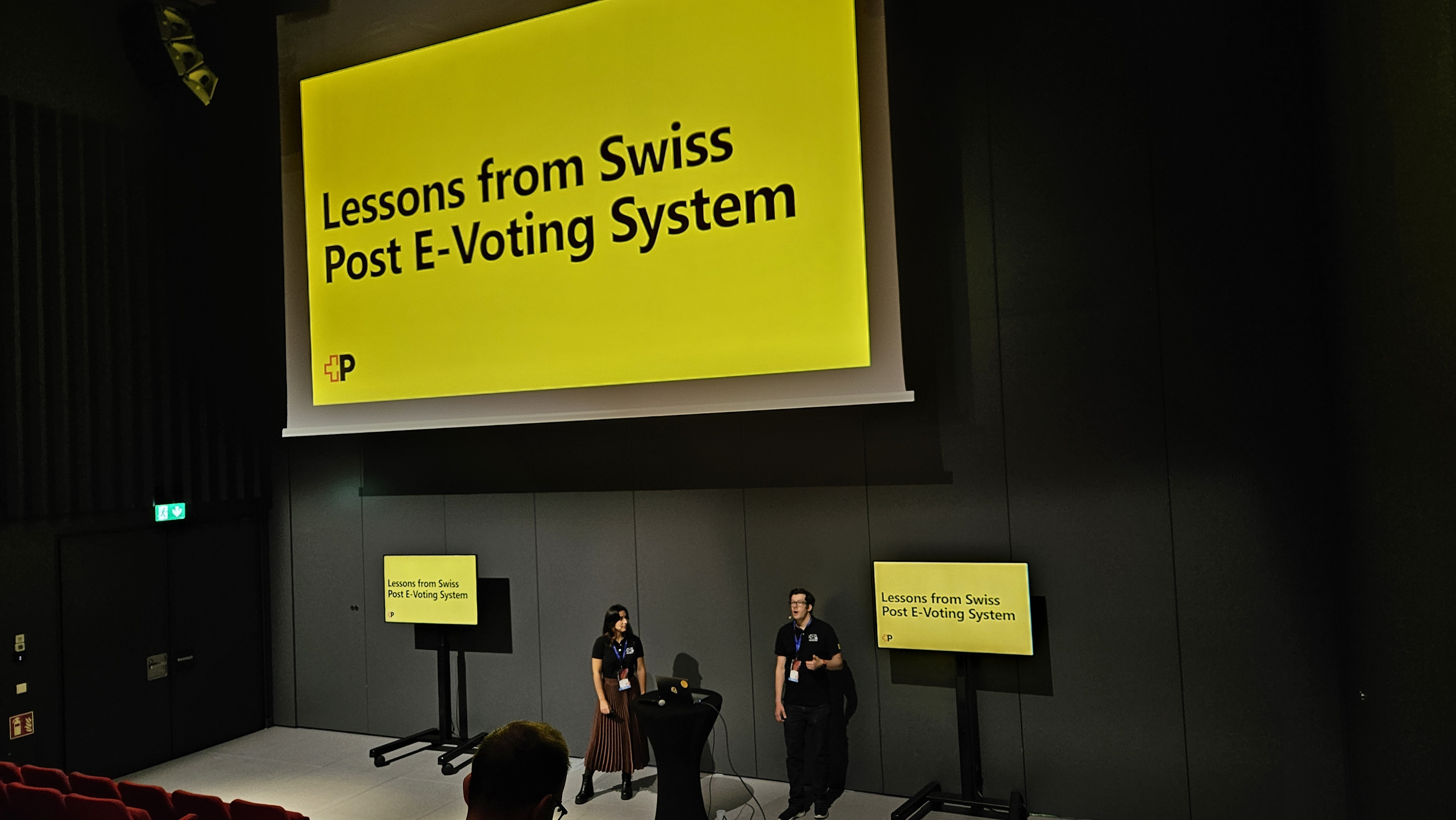 Two people present on a stage in front of a large screen titled “Lessons from Swiss Post E‑Voting System,” with additional smaller displays.