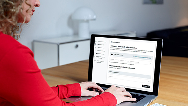 Une personne est assise à une table et utilise un ordinateur portable sur lequel s'affiche un formulaire en ligne. Des documents et un cahier rouge sont posés sur la table. La pièce est lumineuse et meublée de façon moderne.