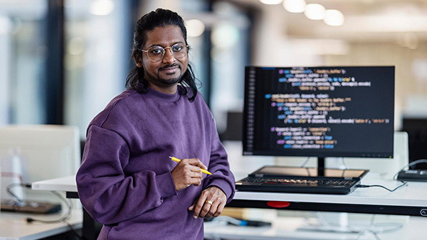 Una persona è in piedi in un ufficio moderno davanti a un monitor su cui è visualizzato il codice di un programma. Indossa un maglione viola, tiene in mano una penna e si appoggia leggermente a una scrivania regolabile in altezza. L'ambiente è luminoso e dotato di attrezzature tecnologiche.