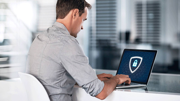 Una persona è seduta alla scrivania e lavora al computer portatile. Sullo schermo è visibile un grande simbolo a forma di cartello con un lucchetto, che rappresenta la sicurezza digitale. L'ambiente appare moderno e aperto, con elementi d'arredo sfocati sullo sfondo.
