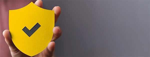 Una mano tiene un simbolo a forma di scudo giallo con un segno di spunta nero. Lo scudo è posto su uno sfondo grigio monocromatico e simboleggia sicurezza o protezione.