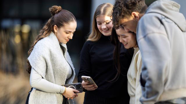 Quattro persone sono in piedi all'aperto, vicine tra loro, e guardano insieme uno smartphone che una di loro tiene in mano. Il gruppo sembra concentrato e immerso in una conversazione o in un'informazione comune. Sullo sfondo si intravedono aree esterne sfocate e strutture edilizie moderne.