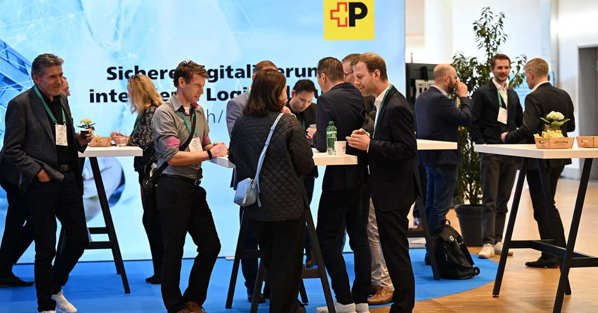 Mehrere Personen stehen und unterhalten sich an Stehtischen in einer Messe- oder Konferenzumgebung. Im Hintergrund ist ein großes blaues Banner mit dem Logo der Post und dem Text „Sichere digitale Lösung“ sichtbar. Auf den Tischen stehen Getränke und kleine Dekorationen. Mehrere Personen stehen und unterhalten sich an Stehtischen in einer Messe- oder Konferenzumgebung. Im Hintergrund ist ein großes blaues Banner mit dem Logo der Post und dem Text „Sichere digitale Lösung“ sichtbar. Auf den Tischen stehen Getränke und kleine Dekorationen.