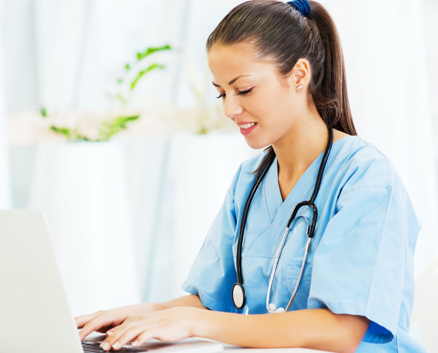 Un'assistente in abiti da lavoro blu con uno stetoscopio al collo lavora su un computer portatile in un ambiente luminoso e accogliente. Un'assistente in abiti da lavoro blu con uno stetoscopio al collo lavora su un computer portatile in un ambiente luminoso e accogliente.