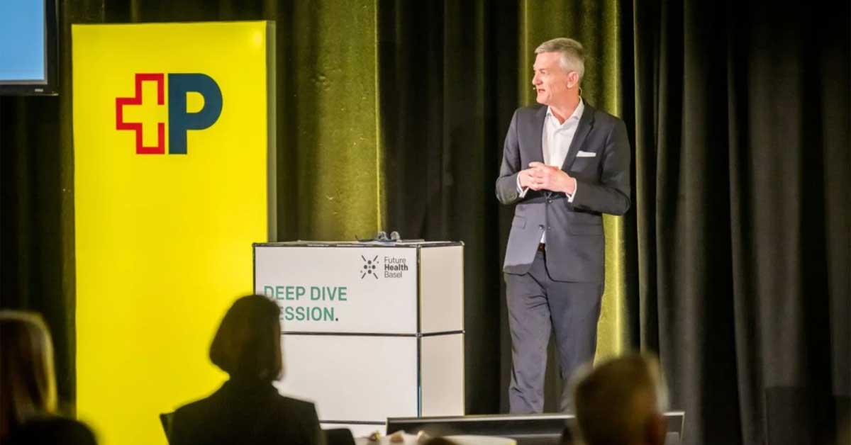 Herr Schneider steht auf einer Bühne neben einem gelben Post-Banner. Vor ihm befindet sich ein Rednerpult mit der Aufschrift „Deep Dive Session“ und dem Logo „Future Health Basel“. Im Vordergrund sind einige Personen im Publikum sichtbar. Herr Schneider steht auf einer Bühne neben einem gelben Post-Banner. Vor ihm befindet sich ein Rednerpult mit der Aufschrift „Deep Dive Session“ und dem Logo „Future Health Basel“. Im Vordergrund sind einige Personen im Publikum sichtbar.