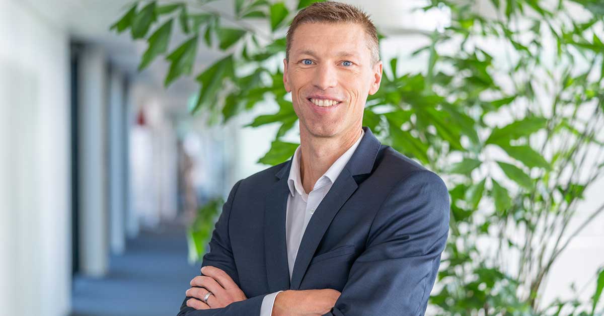 Person wearing a dark blue suit and white shirt stands with arms crossed in a bright, modern hallway. Green plants and blurred architectural details are visible in the background.