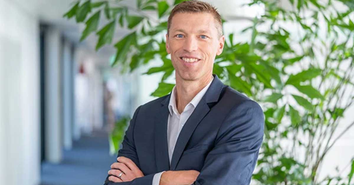 Person in einem dunkelblauen Anzug mit weißem Hemd steht mit verschränkten Armen in einem modernen, hellen Flur. Im Hintergrund sind grüne Pflanzen und unscharfe architektonische Elemente sichtbar. Person in einem dunkelblauen Anzug mit weißem Hemd steht mit verschränkten Armen in einem modernen, hellen Flur. Im Hintergrund sind grüne Pflanzen und unscharfe architektonische Elemente sichtbar.