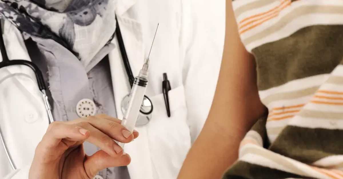 A healthcare professional in a white coat holds a syringe, ready for an injection. Next to them is the arm of a person wearing a short-sleeved striped shirt.