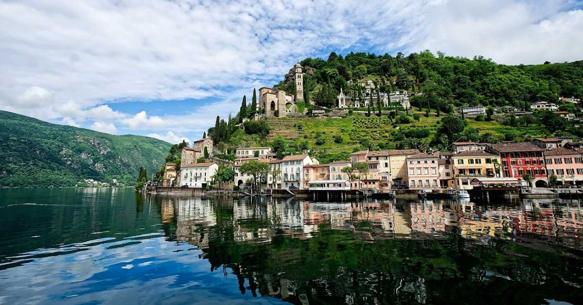 Panoramaaufnahme einer malerischen Ortschaft am Ufer eines Sees im Kanton Tessin. Mehrere farbige Häuser stehen entlang des Wassers, darüber erhebt sich ein grüner Hügel mit einer Kirche und Zypressen. Der See spiegelt die Gebäude und den Himmel mit Wolken wider. Panoramaaufnahme einer malerischen Ortschaft am Ufer eines Sees im Kanton Tessin. Mehrere farbige Häuser stehen entlang des Wassers, darüber erhebt sich ein grüner Hügel mit einer Kirche und Zypressen. Der See spiegelt die Gebäude und den Himmel mit Wolken wider.
