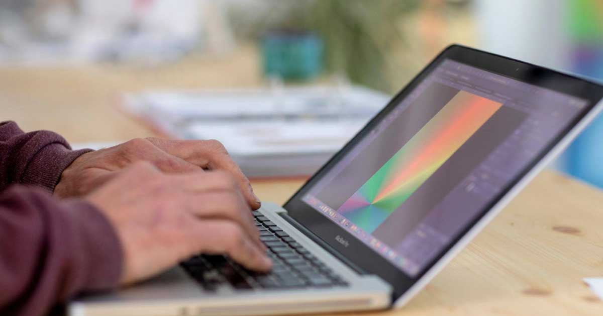 Close-up of hands typing on a laptop keyboard. The screen shows a graphic editing program with a colorful abstract design featuring diagonal shapes in green, red, and blue. The laptop is on a light wooden table, with blurred office items in the background.